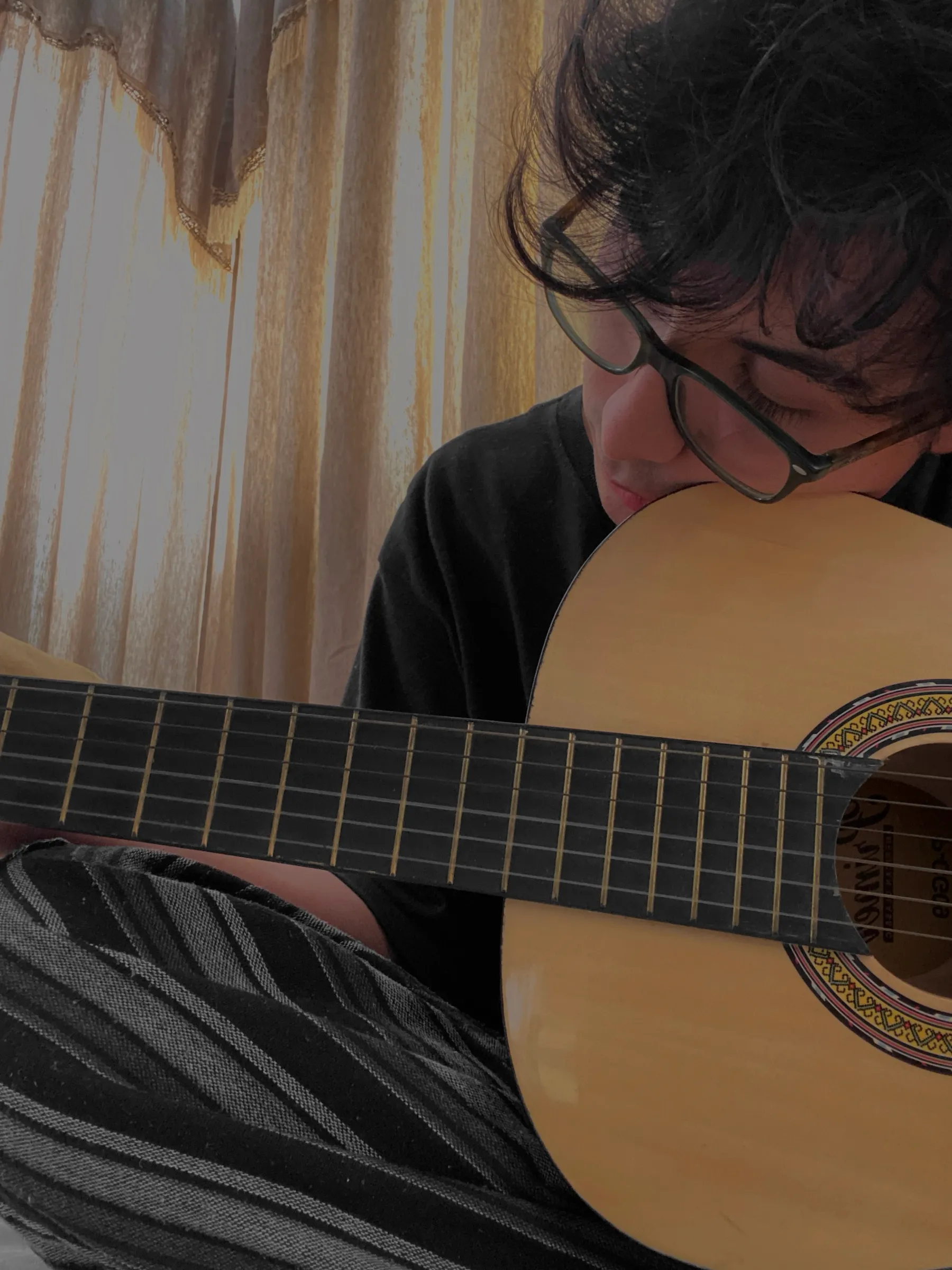 Sebastian playing an acoustic guitar beside a sunlit curtain